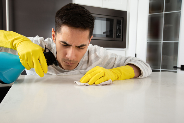 Man wearing yellow gloves cleaning a white quartz countertop with a blue spray bottle and cloth in a modern kitchen, focusing on removing a stain.