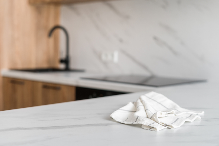 Modern kitchen with a white quartz countertop featuring subtle gray marbling, a neatly placed striped kitchen towel in the foreground, and blurred elements like a black sink, faucet, and cooktop in the background.