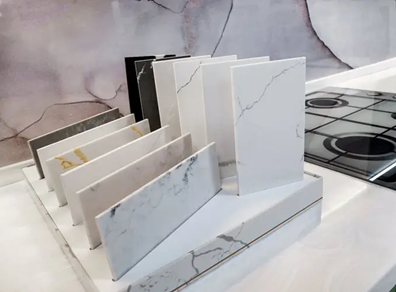 An organized display of marble and stone countertop samples in various colors and patterns, set in a sleek kitchen environment with a glass stove top in the background.