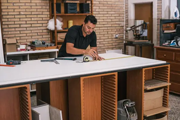 Man measuring a large white countertop in a workshop, surrounded by tools and shelves, showcasing precision in countertop fabrication.
