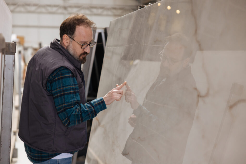 Man inspecting a large, polished natural stone slab with beige and brown veining in a stone warehouse, highlighting the reflective surface and intricate patterns.