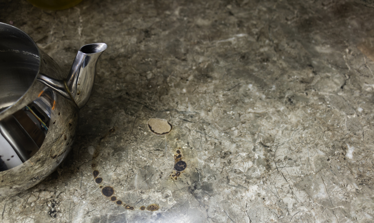 Close-up of a metallic teapot on a granite countertop with visible etching and liquid stains, highlighting the effects of spills on natural stone surfaces.