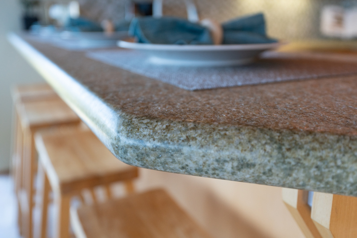 Close-up of a speckled granite countertop with a rounded edge, showcasing its thickness and polished finish, set in a modern kitchen with a blurred table setting in the background